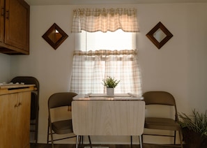 A small dining area with a table and two chairs, a window with plaid curtains, and a wooden cabinet.