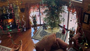 Interior acogedor de una cabaña con un gran árbol de Navidad, paredes de madera, una chimenea y vistas a un paisaje nevado a través de amplias ventanas.