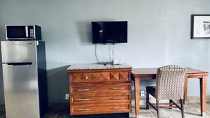 A kitchenette with a microwave, refrigerator, and wooden dresser with drawers. A flat-screen TV is mounted on the wall above a wooden table and chair. - Extended Stay SA (San Antonio)