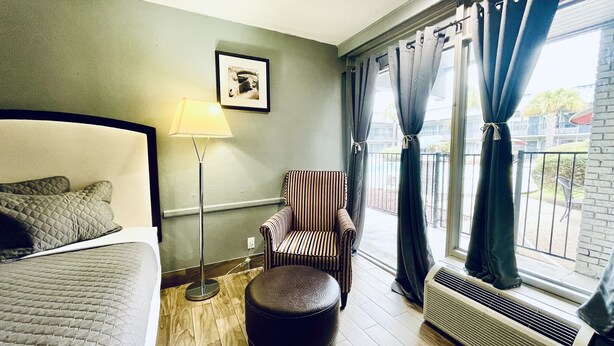 A hotel room with a bed, a standing lamp, a striped armchair, a round ottoman, a window with curtains, and a radiator. - Extended Stay SA (San Antonio)