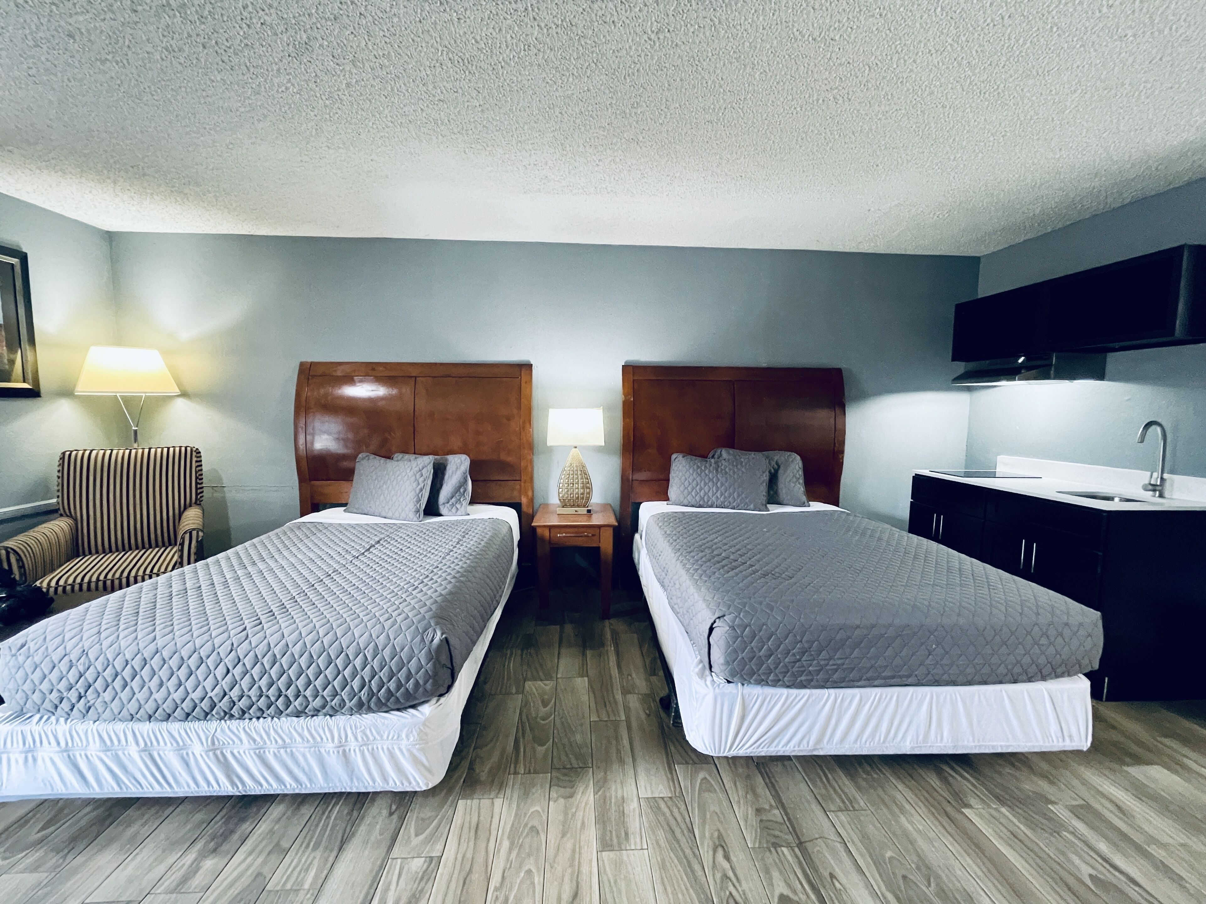 Quarto de hotel com duas camas, uma escrivaninha, uma cadeira, um abajur e uma cozinha compacta.