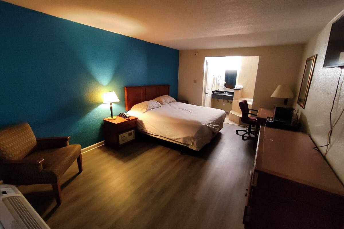 A hotel room with a bed, bedside table with a lamp, a chair, a desk with a chair, and a window.