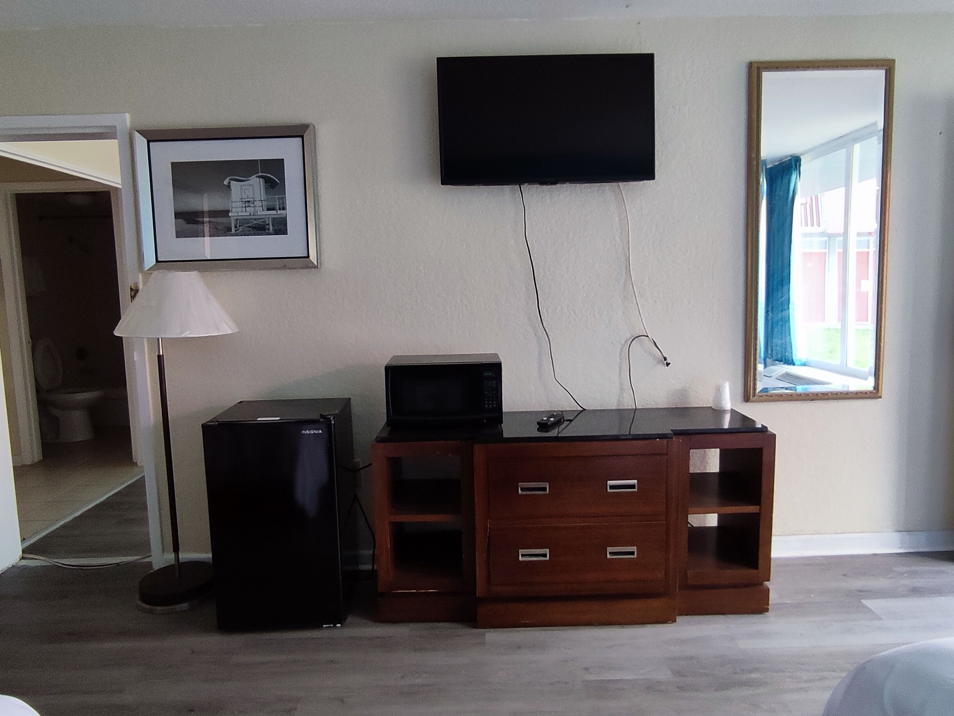 A room with a flat-screen TV mounted on the wall, a wooden entertainment unit with a television and a small refrigerator, a lamp, and a framed picture.