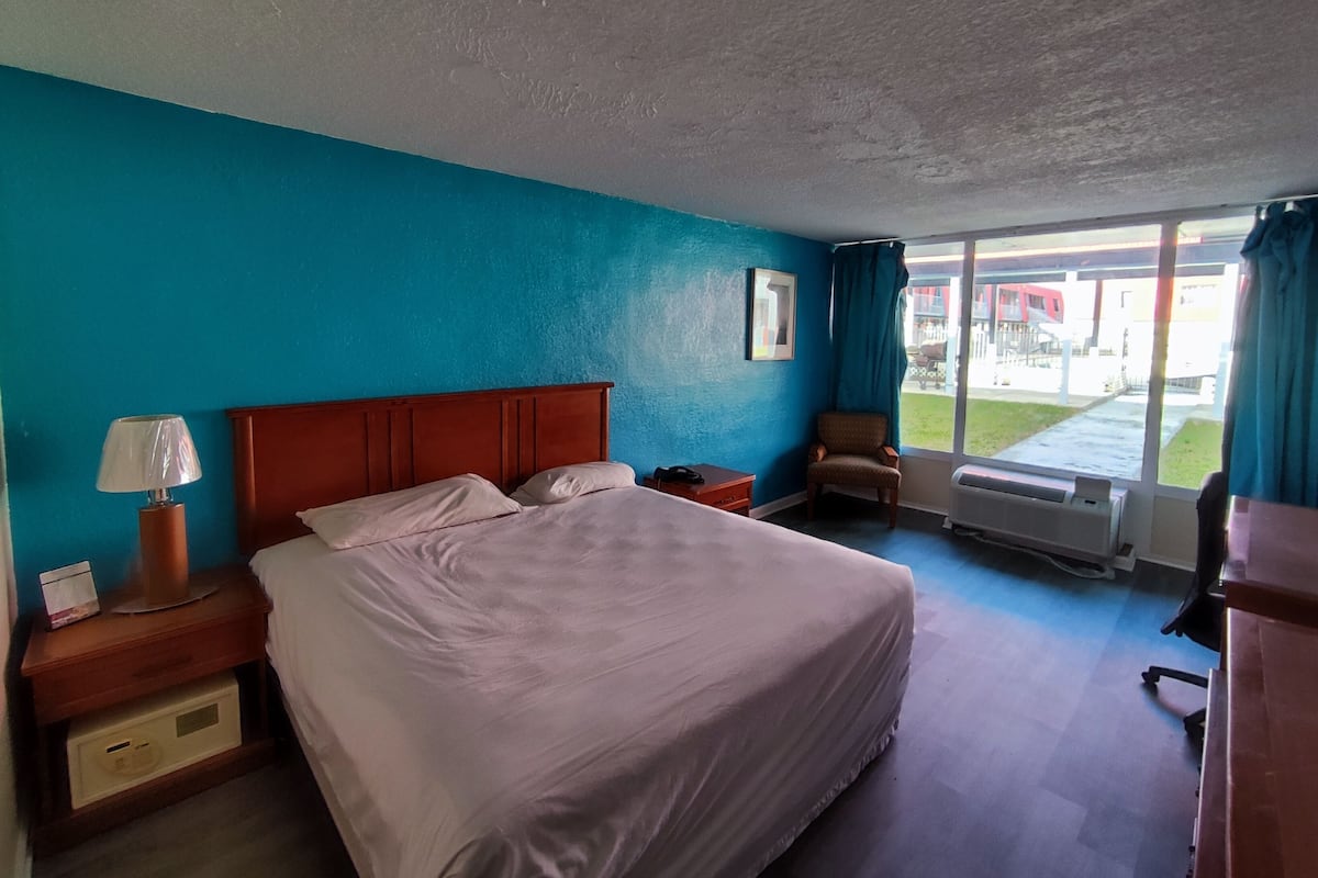 A hotel room with a large bed, a nightstand with a lamp, a chair, and a window with a view of the outside.
