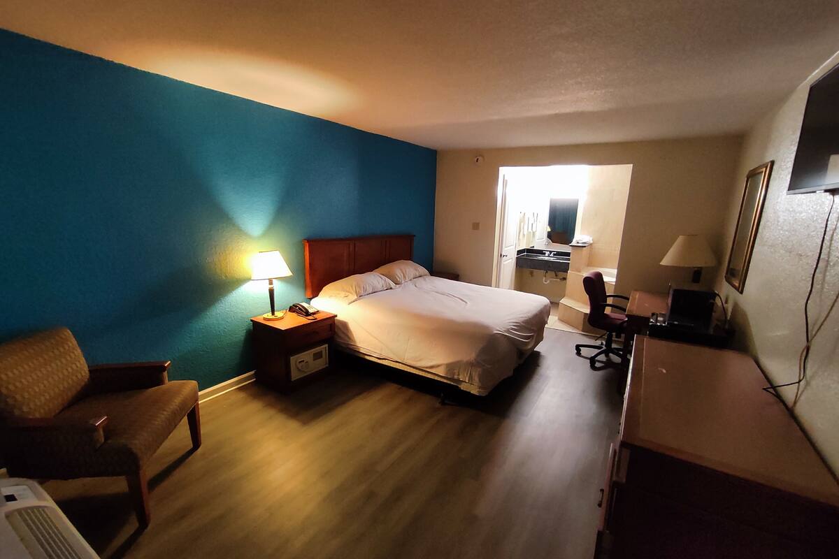 A hotel room with a bed, bedside table with a lamp, a chair, a desk with a lamp, and a mirror.