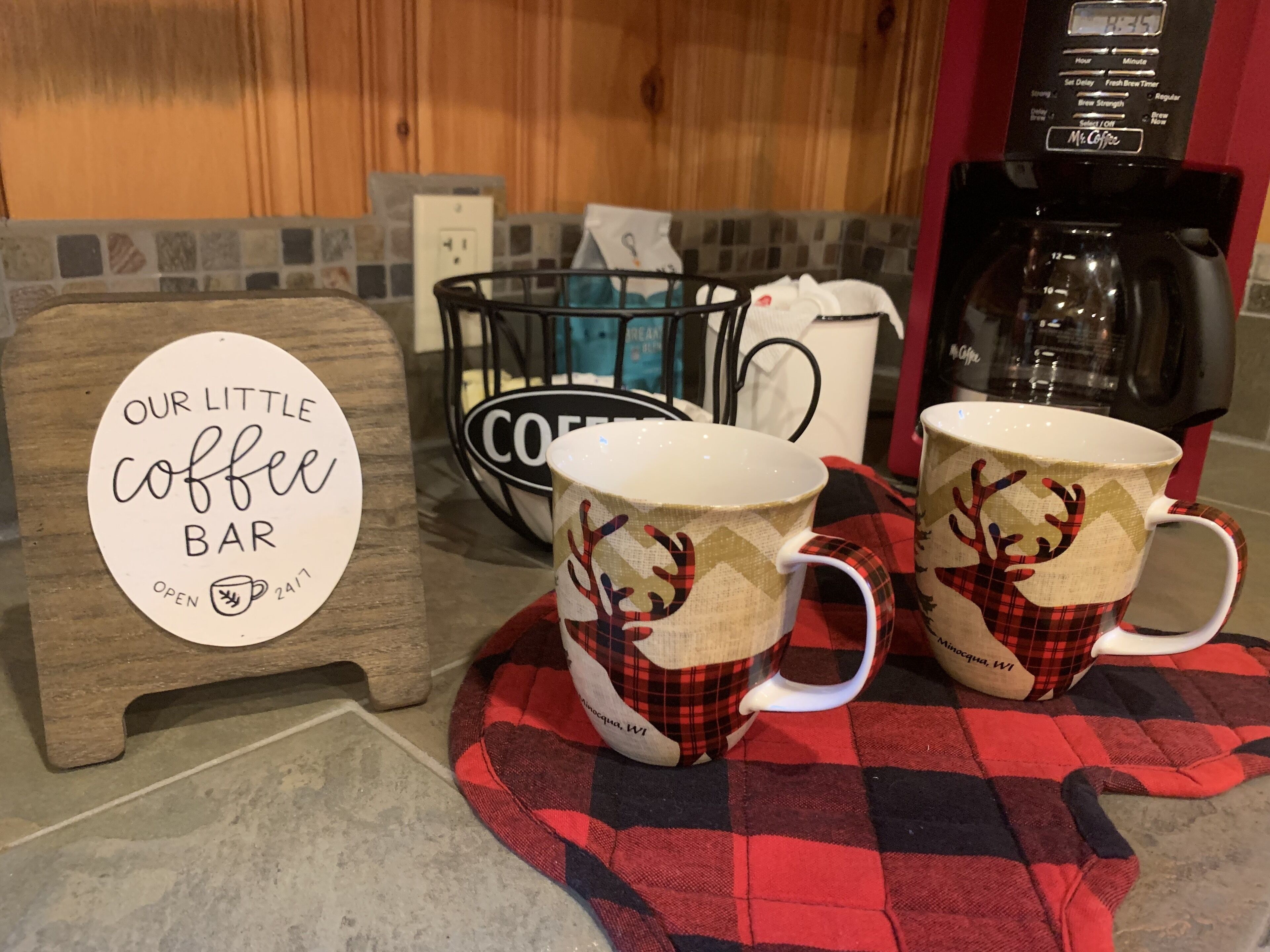 Skilt fra en kaffebar, to kopper med rensdyrmotiver, en kaffemaskine og en kurv med en kaffepose.