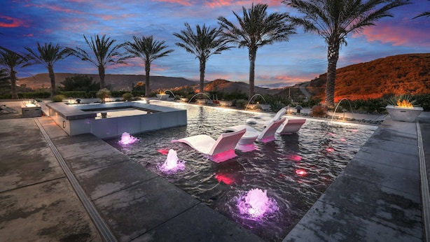 Un espace piscine agrémenté de jeux d’eau et de coins salon, entouré de palmiers et dominé par un paysage montagneux.