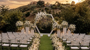 Un lieu de réception en plein air pour un mariage, avec une pelouse verdoyante, des compositions florales blanches et un ciel au coucher du soleil en toile de fond.