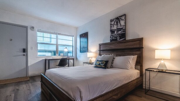 Ein Schlafzimmer mit einem großen Bett, einem Schreibtisch und einem Sessel. Ein Fenster bietet Blick auf Gebäude.