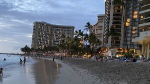 Un hotel frente a la playa con varios pisos, palmeras y gente disfrutando la playa.