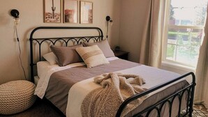 A neatly made bed with a metal frame, a bedside lamp, and a window with curtains. - McKinley House Prime - Patio, Pool and Garage (Amarillo)