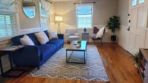 Uma sala de estar com um sofá azul, uma mesa de centro, um abajur e duas janelas com persianas.