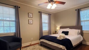 Quarto de hotel com cama, poltrona, ventoinha no teto e duas janelas com cortinas.