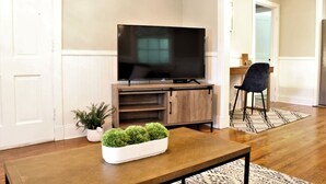 Sala de estar com televisão de tela plana em um suporte de madeira, mesa de centro com uma planta e uma cadeira preta.