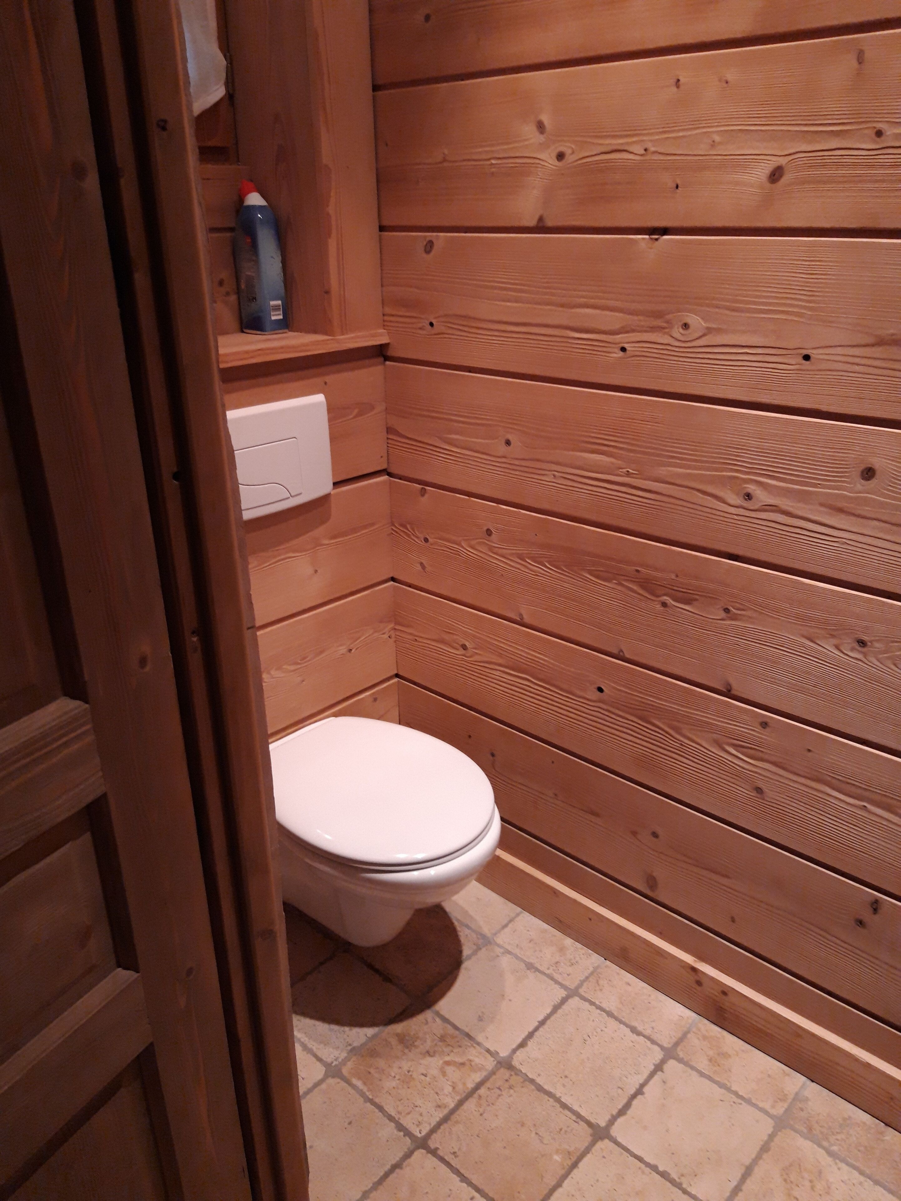 A wooden-paneled bathroom with a white toilet, a shelf with toiletries, and a white electrical outlet.