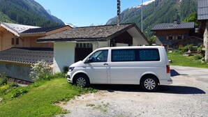 Ein weißer Van parkt vor einem kleinen Gebäude mit einem geneigten Dach, umgeben von Grünpflanzen und Bergen im Hintergrund.