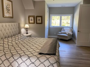 A bedroom with a large bed, a nightstand with a lamp, two framed pictures on the wall, a window with a view of trees, and a chair.