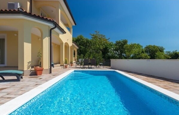 Ein Pool mit Fliesen umgeben, eine Liege und ein Gebäude mit gelber Fassade und Bogenfenstern.