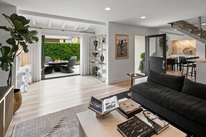 A modern living room with a dark sofa, a coffee table with books, a fireplace, and a view of a dining area and garden.
