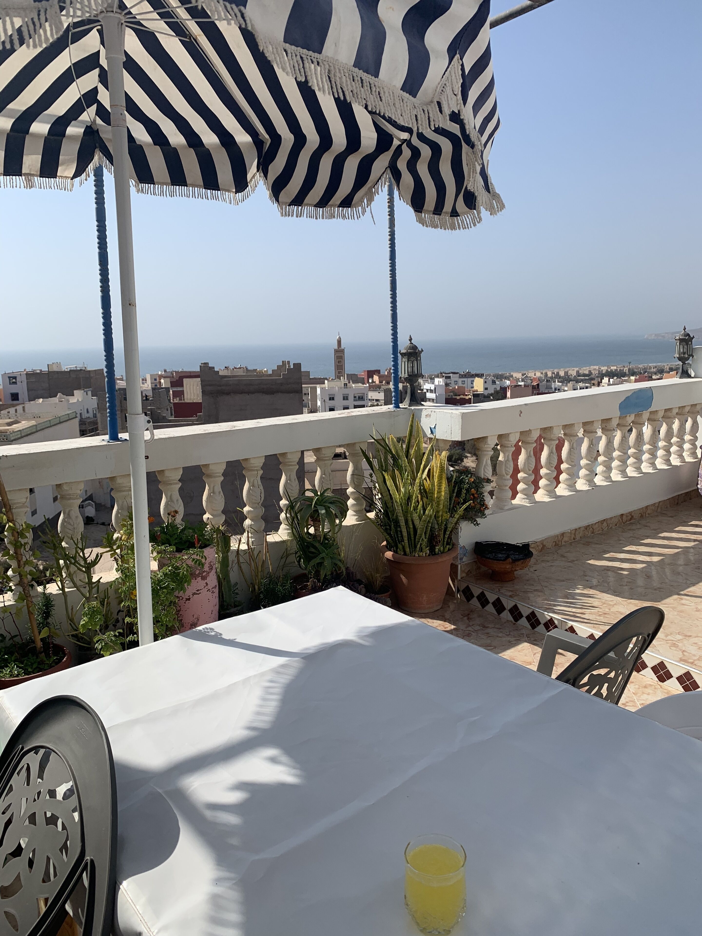 A terrace with a striped umbrella, potted plants, and a table with a glass of drink.