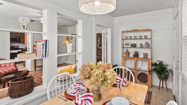 A dining area with a wooden table, white chairs, a chandelier, and a bookshelf with decorative items.