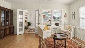 A living room with a wooden floor, a ceiling fan, a white armchair, a sofa, a coffee table, and a patterned rug.