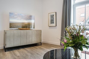 Uma sala de estar moderna com um armário de madeira, uma grande pintura de paisagem, um quadro emoldurado e um vaso com flores.