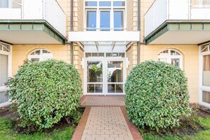 En byggnad med en central entré, två symmetriska, gröna häckar och en gång av tegelstenar som leder fram till entrén.