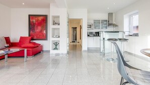 A modern living room with a red sofa, a glass coffee table, and a red abstract painting. It leads to a kitchen with white cabinets and stainless steel appliances.