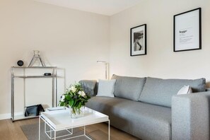 Una sala de estar moderna con un sofá gris, una mesa de centro blanca y una mesita auxiliar de metal con un reloj y un jarrón de flores.