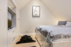 Un dormitorio con techo inclinado, una cama con manta gris, un cuadro en la pared y un tapete mullido negro en el suelo.