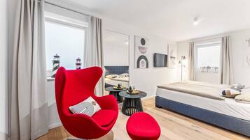 A modern hotel room with a red chair, a small round table, a bed, and a view of a lighthouse.