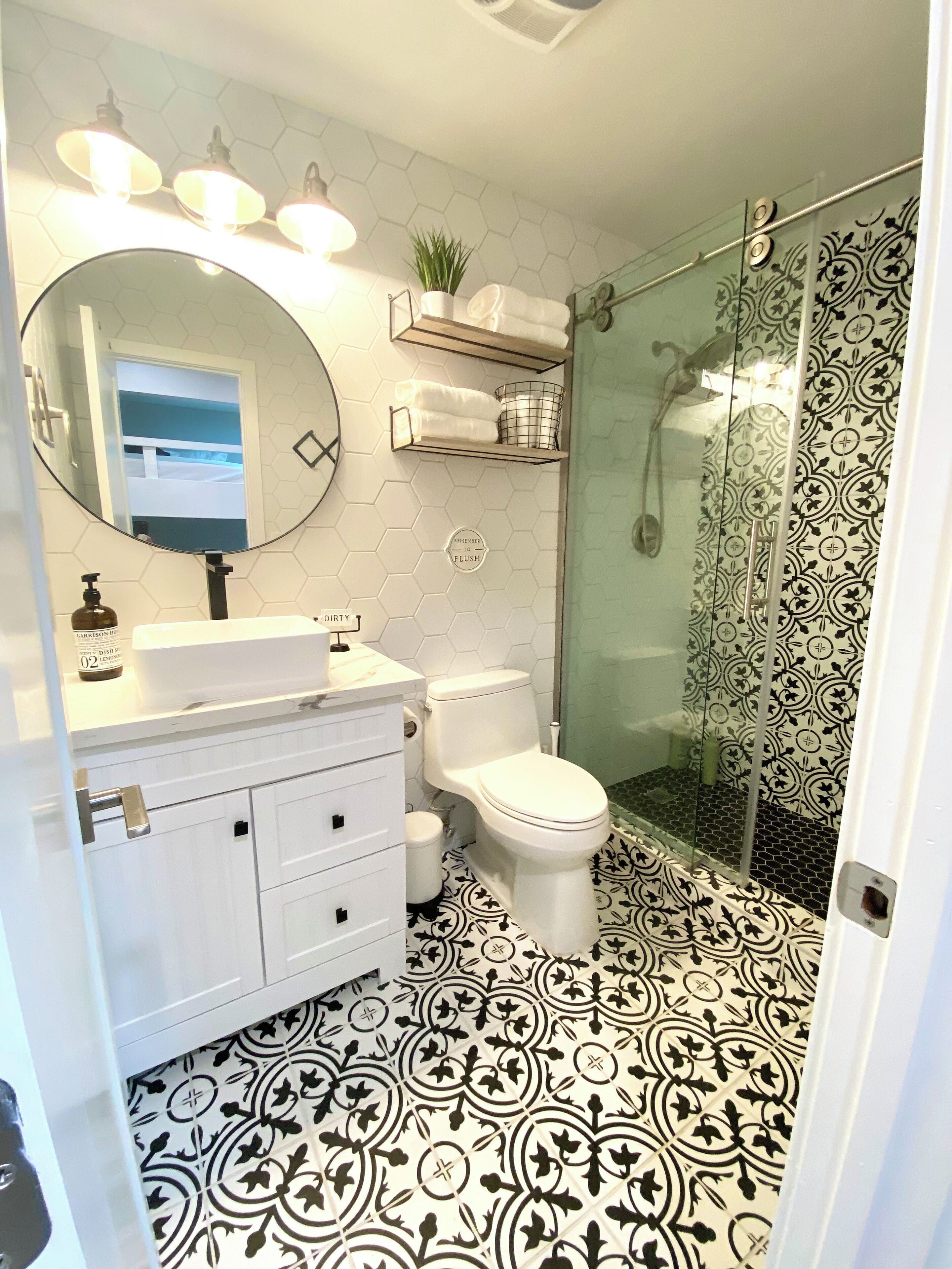 Casa de banho com toucador branco, espelho redondo, sanita e área de chuveiro com azulejos estampados.