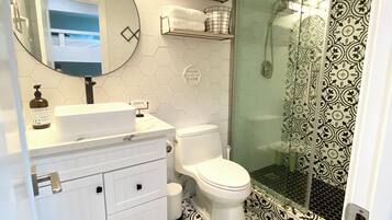 Casa de banho com toucador branco, espelho redondo, sanita e área de chuveiro com azulejos estampados.