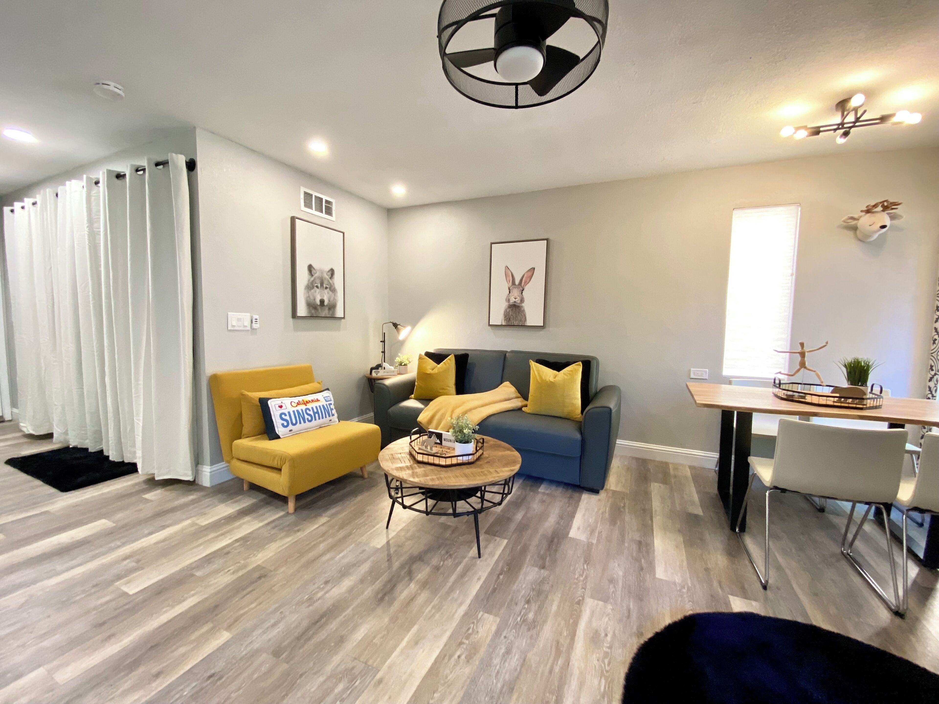 Un salón moderno con un sofá, un sillón y una mesa de centro. Hay dos cuadros enmarcados en la pared, un ventilador de techo y una ventana con cortinas.