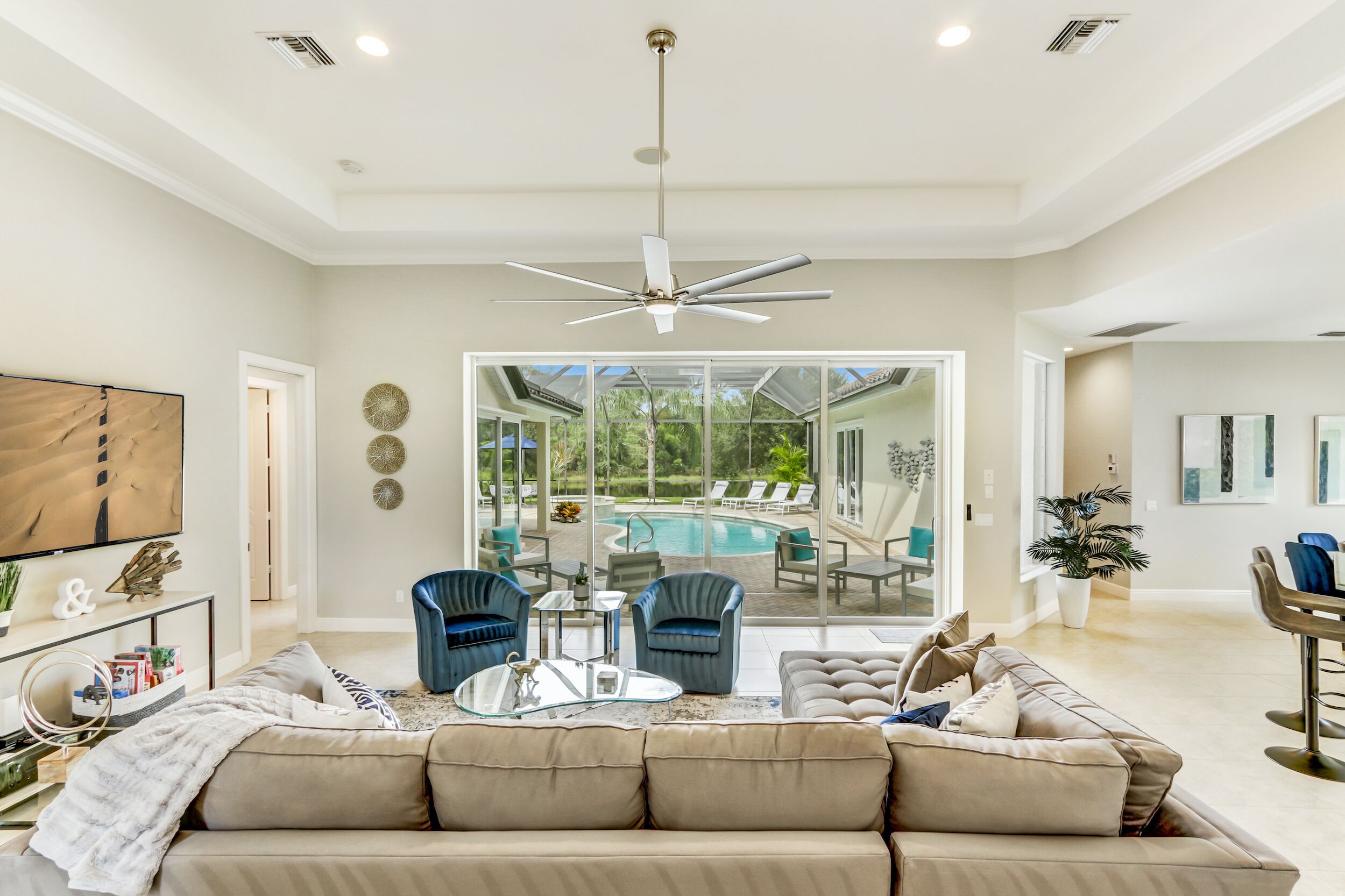 A spacious living room with a large sofa, two armchairs, a coffee table, and a ceiling fan. A television is mounted on the wall, and there is a large window providing a view of a swimming pool and outdoor seating area.