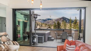 A modern living room with a large glass door leading to an outdoor patio. Inside, there is a leather sofa, a wooden coffee table, and a unique chair with horn-like legs. The patio features outdoor furniture, a hot tub, and a view of mountains.