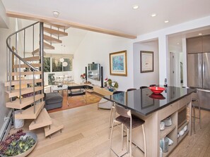 Una cocina moderna con una isla central, un comedor con sillas, una escalera y una sala de estar con sofá y mesa de centro.