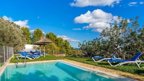 Une piscine rectangulaire à l’eau d’un bleu limpide, entourée d’une terrasse carrelée de couleur blanche. On y trouve des chaises longues bleues et un gazebo à toit de chaume.
