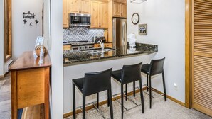 A modern kitchen with wooden cabinets, stainless steel appliances, and a granite countertop. There are three black bar stools and a wooden dining table. - Millrace 6-A | Cozy 2BR Condo, Creek Views, Vail Cascade (Vail)