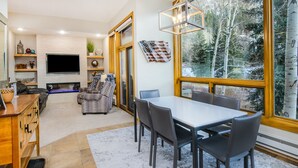 A modern living room with a fireplace, a dining area with a large window, and a TV mounted on the wall. - Millrace 6-A | Cozy 2BR Condo, Creek Views, Vail Cascade (Vail)