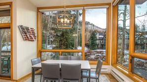 A dining area with a table and chairs, large windows with a view of a snowy landscape, and a chandelier. - Millrace 6-A | Cozy 2BR Condo, Creek Views, Vail Cascade (Vail)