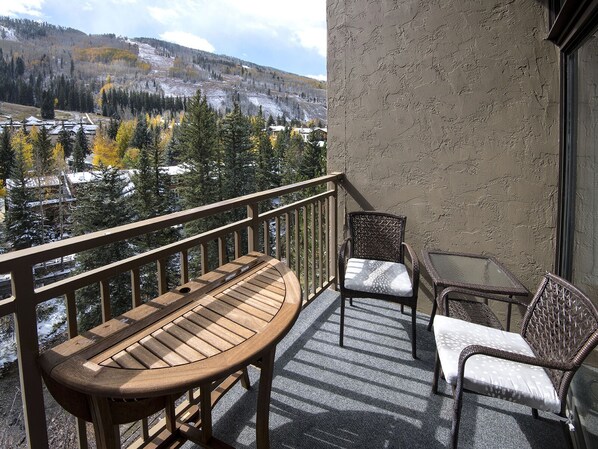A balcony with a wooden table and two wicker chairs, overlooking a mountainous landscape with autumn-colored trees and snow patches. - Wren at Vail | 1BR+Loft Condo, Steps from Lifts, Pool (Vail)