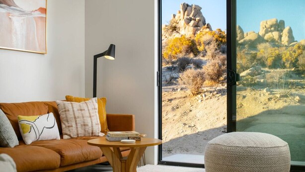 A modern living room with a leather sofa, a round ottoman, a small wooden table, and a floor lamp. A large window offers a view of rocky terrain and sparse vegetation.