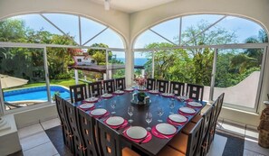 A dining area with a large table set for 12, surrounded by chairs, with a view of a swimming pool and lush greenery. - Oceanview FAST WiFi Private Yard Pool Executive Chef Staff Amazing Views (boca de tomatlan)