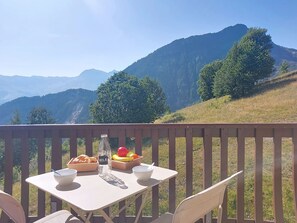 Ein Tisch mit einer Flasche, Schalen und einer Obstschale auf einem Balkon mit Blick auf Berge.
