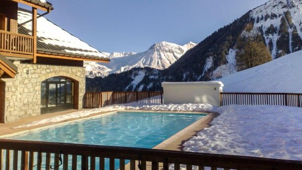 Zona de piscina con un edificio de piedra, terraza de madera y vistas a montañas nevadas.
