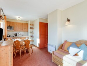 Un espacio habitable compacto con kitchenette, una zona de comedor con sillas de madera, un sofá con cojines y una puerta roja.