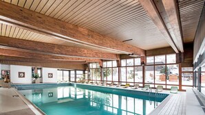 Ein großer, überdachter Schwimmbereich mit Holzbalken, großen Fenstern und Liegestühlen am Beckenrand.