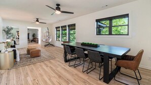 Un vaste espace de vie comprenant une grande table à manger, de nombreuses chaises et des ventilateurs de plafond. De grandes fenêtres laissent entrer la lumière naturelle et offrent une vue sur la verdure environnante.